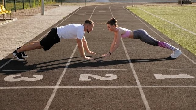 Sports active man and women train in the summer outdoors in a public stadium, they stand in the plank and slap each other's hands.