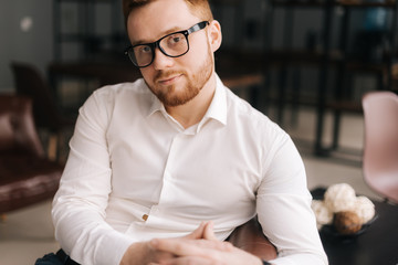 Creative young business man wearing stylish glasses in trendy clothing looking at camera while sitting indoors on chair in modern office. Portrait of bearded handsome gentleman wearing white shirt.