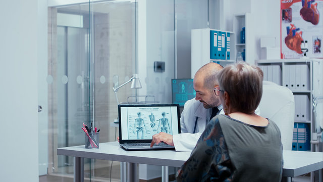 Explaining human skeleton to elderly patient from a booklet on laptop. Radiology and radiography in modern private hospital or clinic with medical staff walking in background, nurse working