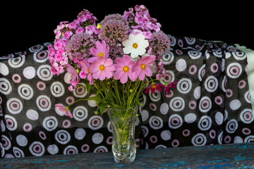 Fototapeta premium On an old wooden bench in a crystal vase against a bright background of fabric, there is a bouquet of cosmos and phlox flowers.
