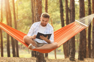 Young man with book relaxing in hammock outdoors