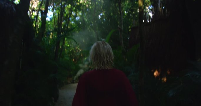 Woman with blonde hair and red dress walks along nature path surrounded by a deep green jungle forest and palm leaves while on vacation