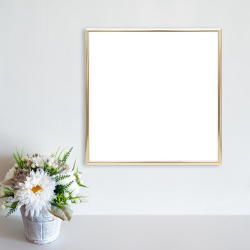Gold frame mockup on white wall with pot of flowers on white surface. Copy space.