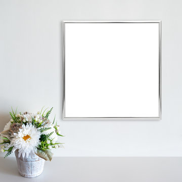 Silver frame mockup on white wall with pot of flowers on white surface. Copy space.