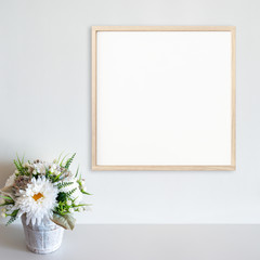 Wood frame mockup on white wall with pot of flowers on white surface. Light wood. Copy space.