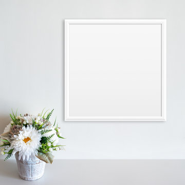 White frame mockup on white wall with pot of flowers on white surface. Copy space.
