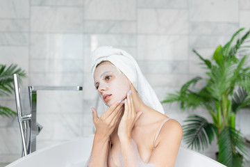 Woman wearing towel on head applying  face sheet mask while taking bath with soap foam.