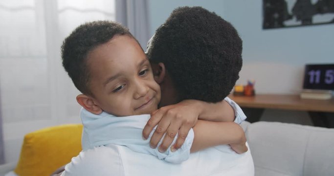 Close up portrait of cute little African American child hugging his father at home. Rear of man hugs happy small boy in living room indoors. Lovely family relations. Happiness concept