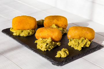 Croquettes fried in oil, with guacamole sauce on black stone plate at sunny kitchen. Side view on white wooden background. Traditional spanish snack