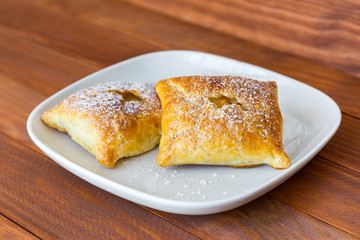 Freshly baked sweet buns sprinkled with powdered sugar and cinnamon on white plate on wooden table, side view, close-up.