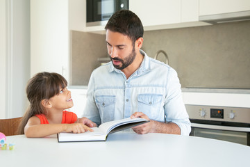 Handsome dad discussing book with daughter in kitchen. Cute girl in red shirt pointing with finger in text, sitting and learning with young Latin father. Family time, childhood and reading concept