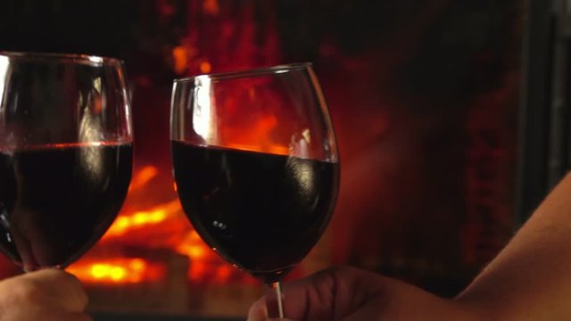 Close-up of the hands clinking glasses with red wine on the background of a burning fireplace