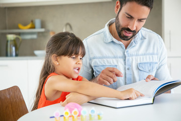Lovely cute girl pointing at text and talking with bearded dad. Handsome young father teaching...