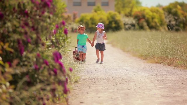 The cute children in hats are walking slowly together along the road with a pink suitcase. Summer travels and adventures
