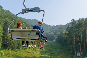 People go to ski elevator in Krasnoyarsk park. View from the back. Summer, green forest, blue sky,...