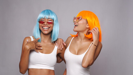 Portrait of two beautiful happy Asian women friends in bright blue and orange wigs and sunglasses...