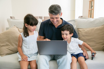 Two happy kids and their dad using laptop while sitting on couch at home, staring at display. Medium shot, front view. Communication or leisure at home concept