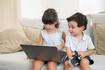 Cute little brother and sister sitting on couch at home and using laptop, watching video, cartoon...