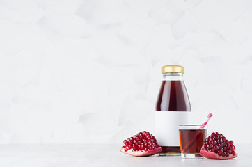 Bright red pomegranate juice in glass bottle mock up with blank label, straw, wine glass, fruit grains on white wood table in light interior, template for packaging, advertising.