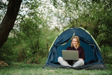 Woman working on laptop in tent in nature. Young freelancer sitting in camp. Relaxing in camping site in forest, meadow. Remote work, outdoor activity in summer. Happy girl relaxing, work on vacation.