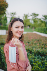 Fototapeta premium Young woman outdoor holding medical face mask in hand and smiling. Afternoon in the park. Selective focus.