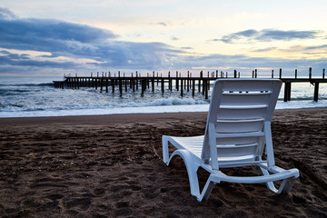 Sunbed on the sand beach and view to water of sea, waves with white foam, pierce and sky with clouds in a nice evening.