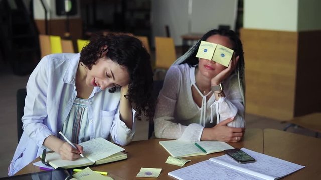 Two caucasian women making fun during their preparation for exams. Funny girl with dreadlocks sitting having sticker yellow papers with painted eyes while her female friend is writing her notebook