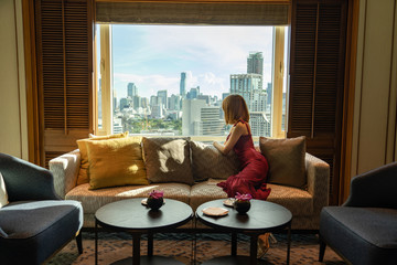 Asian beautiful woman in a red dress, sitting on a sofa chair, pouring out a window with a clear view of the capital's skyscrapers in the morning..