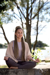 A woman does yoga asanas, meditates in nature. Young girl with afro-braid in sportswear in a lotus position with a defocused background. Healthy and Yoga Concept.