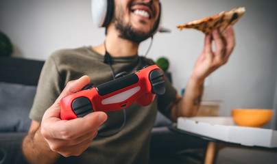 Happy man playing online video games while eating pizza fast food - Young gamer having fun on new technology console - Gaming entertainment and youth millennial generation lifestyle concept
