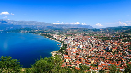 City buildings with red roofs at the shore of a lake with mountains in the background. View of...