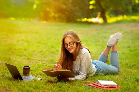 Attractive Young Girl In Eyeglasses Reading Book Outdoors, Enjoying Studying In Park