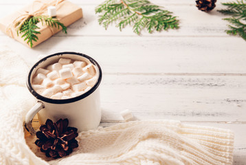 cocoa mug with marshmallows, white sweater, cones, gift box, green branches of thuja, fir branches on a white wooden background, hot winter drink