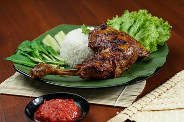 Tasty grilled juicy chicken with golden brown skin served with plain white rice, chili sauce and various vegetables. with a beautiful bamboo plate on a wooden table.