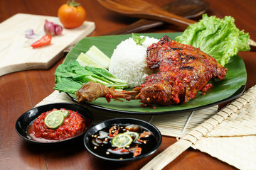 Tasty grilled juicy chicken with golden brown skin served with plain white rice, chili sauce and various vegetables. with a beautiful bamboo plate on a wooden table.