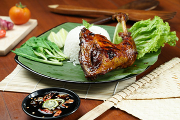 Tasty spicy grilled juicy chicken with golden red skin served with white rice, chili sauce and various vegetables. with a beautiful bamboo plate on a wooden table.