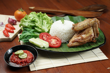 Tasty fried juicy chicken with golden brown skin served with plain white rice, chili sauce and various vegetables. with a beautiful bamboo plate on a wooden table.
