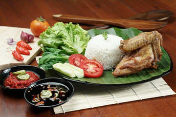 Tasty fried juicy chicken with golden brown skin served with plain white rice, chili sauce and various vegetables. with a beautiful bamboo plate on a wooden table.