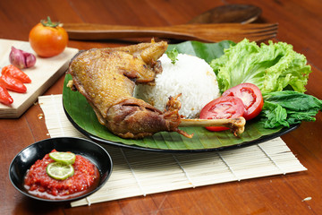 Tasty fried juicy chicken with golden brown skin served with plain white rice, chili sauce and various vegetables. with a beautiful bamboo plate on a wooden table.