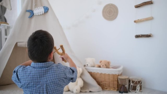 Back view of little boy shooting with slingshot at paper target on room's wall