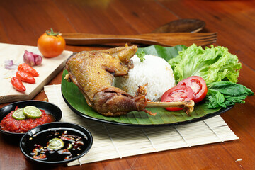 Tasty fried juicy chicken with golden brown skin served with plain white rice, chili sauce and various vegetables. with a beautiful bamboo plate on a wooden table.