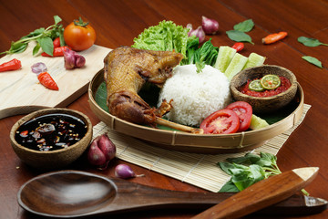 Tasty fried juicy chicken with golden brown skin served with plain white rice, chili sauce and various vegetables. with a beautiful bamboo plate on a wooden table.