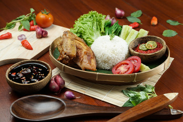 Tasty fried juicy chicken with golden brown skin served with plain white rice, chili sauce and various vegetables. with a beautiful bamboo plate on a wooden table.