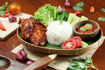Tasty spicy grilled juicy chicken with golden red skin served with white rice, chili sauce and various vegetables. with a beautiful bamboo plate on a wooden table.