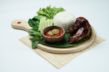 Grilled chicken with brownish skin laid out on top of a wooden sheet on a white background, plus white rice, chili sauce and various vegetables, view from above. commonly used for menus and box wrap i