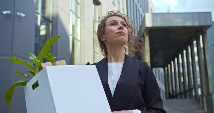 Young woman worker in a formal suit walking around the business district, viewing the scyscapers. Beautiful curly haired accountant holding a box with a job stuff outdoors. Hiring, traineeship.