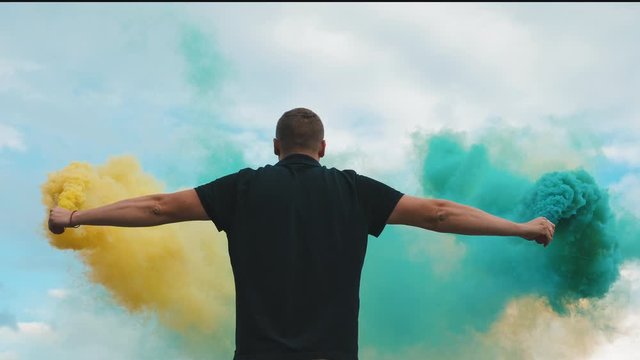 Back view of a man in a black T-shirt with a yellow and blue smoke bomb in his hands in slow motion