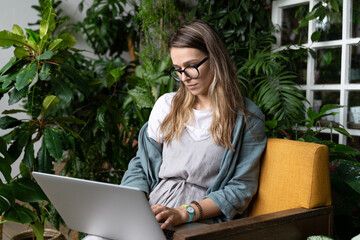 Woman gardener wear linen dress, sitting on chair in green house, working on laptop surrounded by...