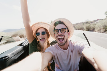 Happy excited couple taking a selfie driving on a convertible car at holidays. Young people at vacation on a rent car.