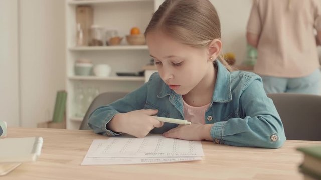 Medium shot of blond-haired Caucasian schoolgirl is sitting at table in the kitchen and doing homework while her mother cooking on background